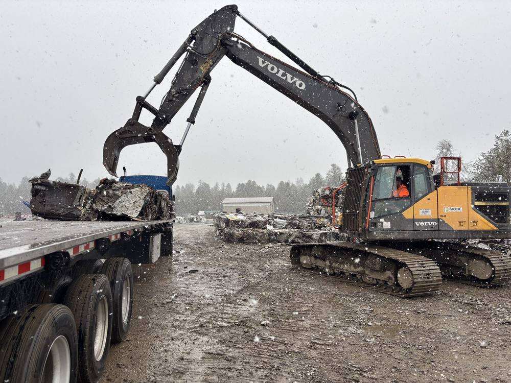 A Volvo excavator with a grapple loading scrap onto a flatbed trailer during a snowstorm