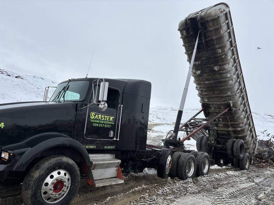 A black Sarsten Kenworth with its end dump trailer raised, unloading on a snowy job site